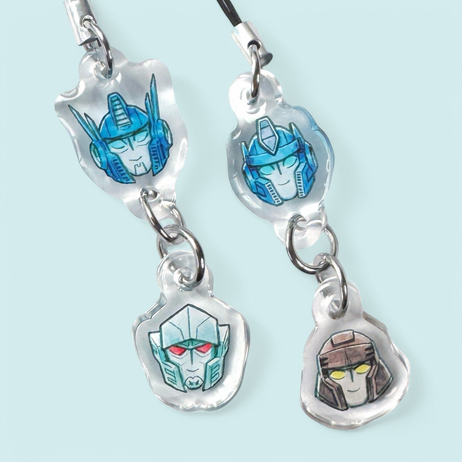 Godliath acrylic phone charms featuring hand-drawn robot character heads in blue, grey, and brown on clear pendants.