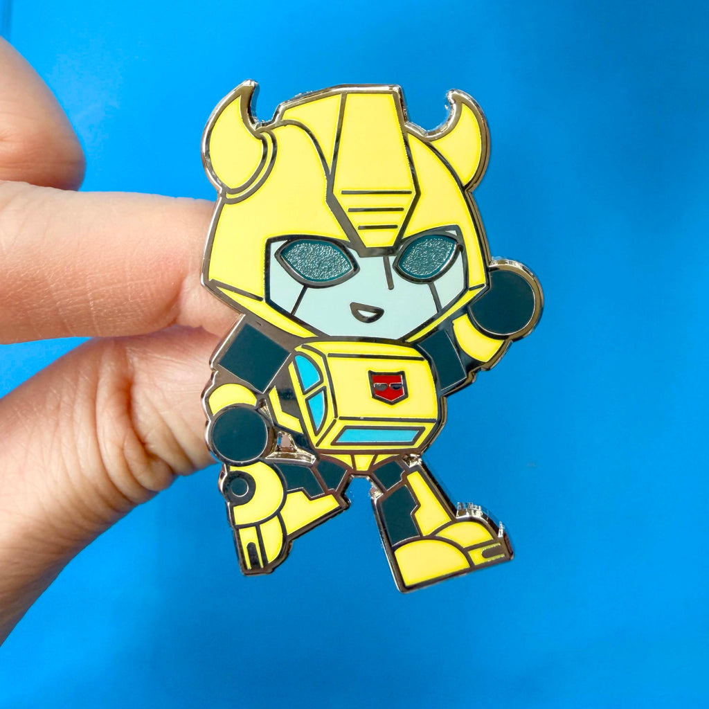 A hand holding a chibi Transformers Bumblebee pin against a bright blue background.