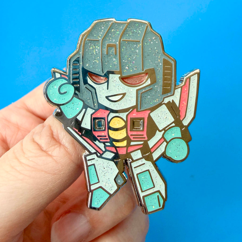 A hand holding a chibi Transformers Starscream enamel pin in his ghost form. The pin has full glitter enamel.