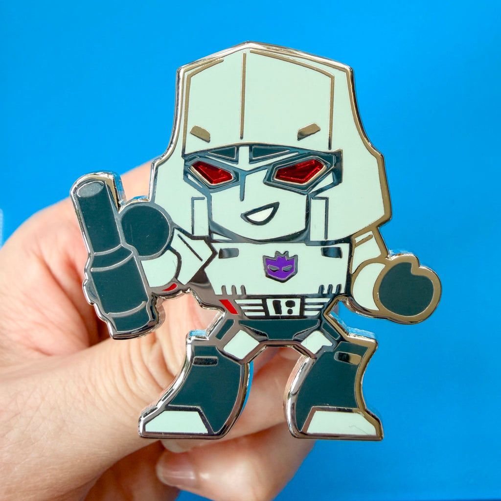 A hand holding a Transformers Megatron hard enamel pin against a bright blue background.