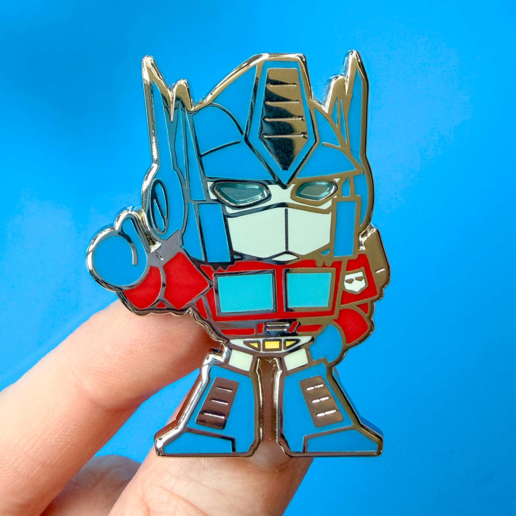 A close-up of a hand holding an Optimus Prime enamel pin against a bright blue background.