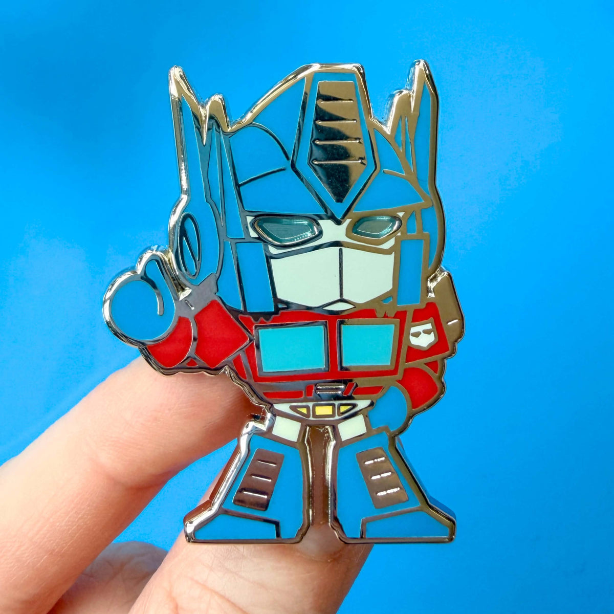A close-up of a hand holding an Optimus Prime enamel pin against a bright blue background.