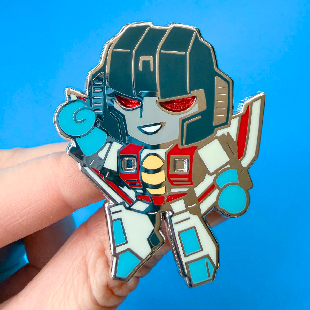 A chibi Transformers Starscream pin held by a hand against a solid blue background. 